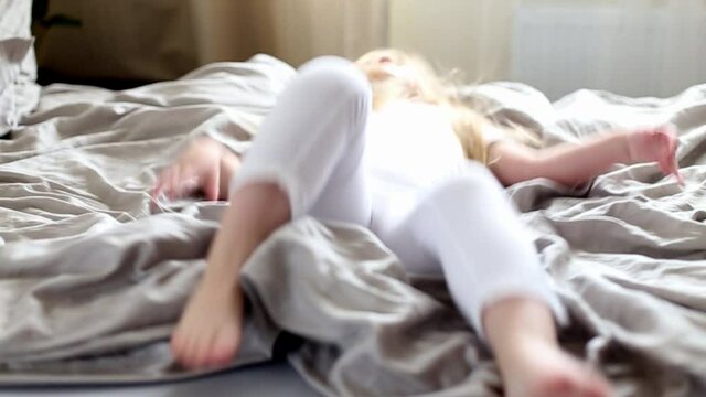 Happy little girl jumping on a grey bed and falling in the morning. childs feet on blanket close. slow motion