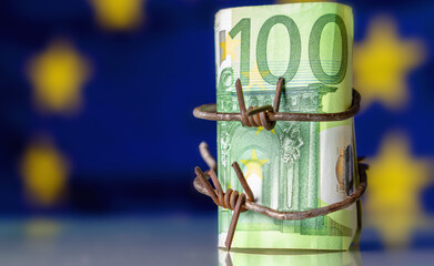 The global economic recession and crisis  of European Union. One hundred EURO bancnotes wrapped in barbed wire against flag of EU. Copy space for design. Horizontal image.