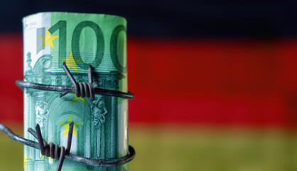 One hundred EURO bancnotes wrapped in barbed wire against flag of Germany as symbol of global economic crisis and recession. Selective focus on money. Copy space. Horizontal image.