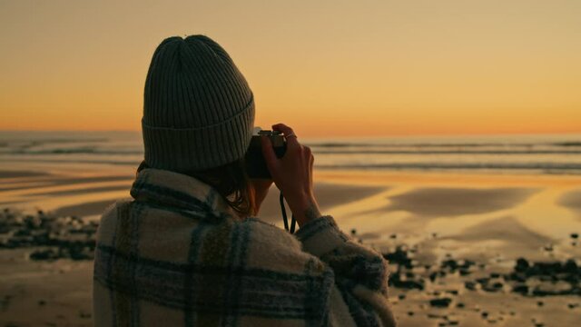 Cinematic and beautiful shot in golden yellow sunset colours of young millennial woman make photos of beach at sunset. Hipster photographer create content. Authentic real person at epic location