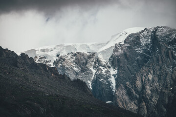 Dark atmospheric surreal landscape with dark rocky mountain top in low clouds in gray cloudy sky. Gray low cloud on high pinnacle. High black rock with snow in low clouds. Surrealist gloomy mountains.