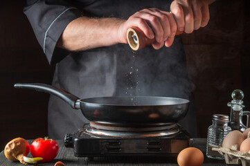 The chef adds pepper while cooking eggs in pan. Work environment on vintage kitchen table