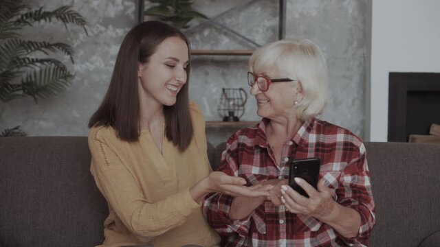Close-up portrait of two nice attractive lovely charming friendly focused women granddaughter teaching grandmother using digital device in light white interior room house flat apartment