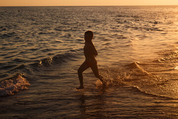 Silhouette of boy running barefoot along the shoreline on the beach at sunset in the summer
