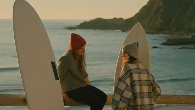 Two young female surfers stand over sunset beach and discuss surfing conditions and waves. Millennial hobby concept. Active outdoor lifestyle wanderlust