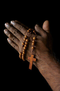 Praying hand of an old Indian Catholic man with wooden rosary isolated on a plain black background.