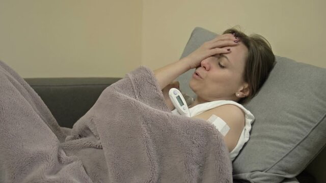 Young woman lies on the sofa, wrapped in a plaid. She has flu or cold symptoms and a very high fever. Side effects of the coronavirus vaccines.