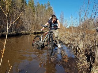 Girl cyclist overcomes wading deep puddles on clay forest road in spring