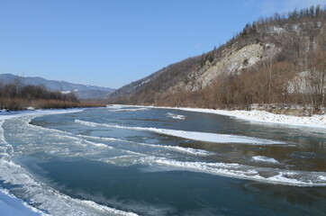 A frozen river in a wintry landscape. Winter landscape with forest, cloudy sky and sun.