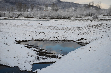 A frozen river in a wintry landscape. Winter landscape with forest, cloudy sky and sun.