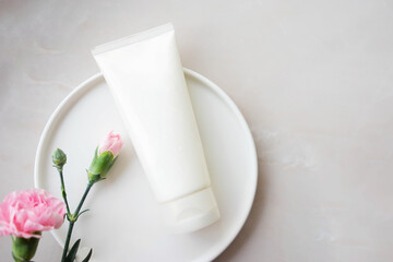 white cosmetic tube with face cream, body lotion or cleanser on a plate with pink flowers on a light background. Sensitive skin care concept. Copy space