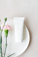white cosmetic tube with face cream, body lotion or cleanser on a plate with pink flowers on a light background. Sensitive skin care concept. Vertical