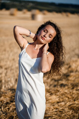 Portrait of a young curly-haired woman in a wheat field, where wheat is mowed and sheaves are standing, enjoying nature. Nature. sun rays Agriculture