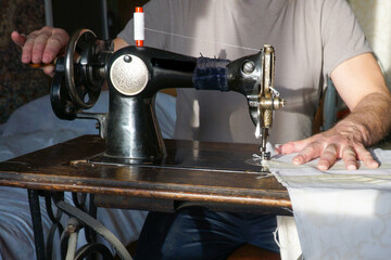   A man sews on an old sewing machine, holding the fabric with one hand and twisting the drive...