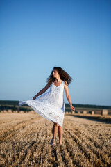 A young beautiful woman in a white summer dress stands on a mown wheat field with huge sheaves of hay, enjoying nature. Nature in the village. Selective focus