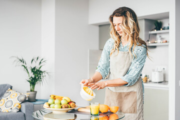 Pretty curly haired lady in grey apron pours fresh orange juice into cup from squeezer at glass table in light contemporary kitchen in morning