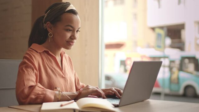 Medium shot of young pretty mixed-race female freelancer wearing trendy casual clothes and accessories sitting at cafe table, working on laptop and making notes in notebook