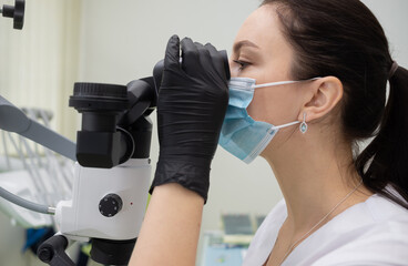 A woman dentist with dark hair in a mask and gloves looks into a dental instrument - a microscope in the office of the clinic. Medicine, dentistry concept.