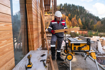 Man builds a cottage on the background of a beautiful mountain. Carpenter on the country house construction. Sawing machine next to the house under construction. Man is cutting boards on a machine.
