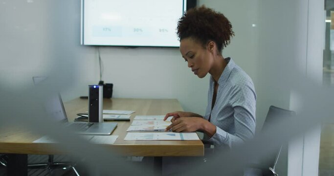 Mixed race businesswoman sitting at desk, checking documents and using laptop in office