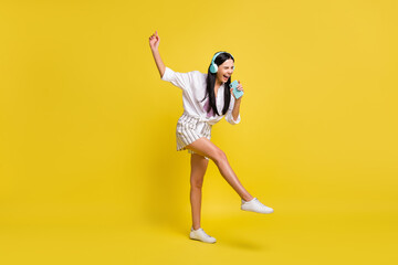 Full size photo of young cheerful smiling positive girl in headphones singing in phone isolated on yellow color background