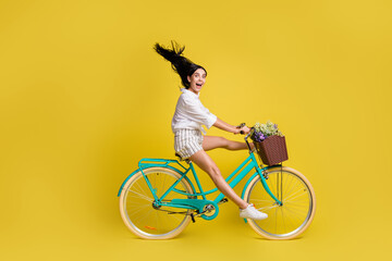 Full size profile side photo of young charming lovely girl with flying hair riding bicycle isolated on yellow color background