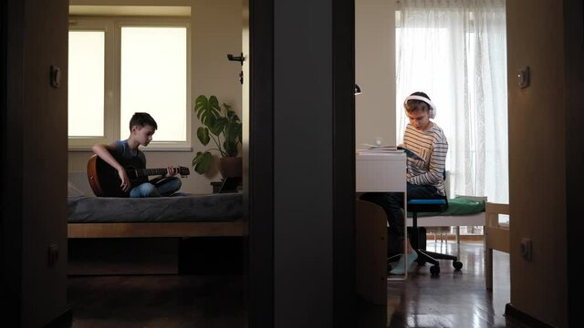 Teenager studying online using laptop computer. Brother learning to play guitar using tablet in bedroom. View through open doors to two rooms from hallway. Online education, entertainment at home
