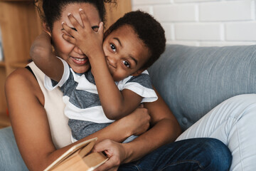 Black smiling mother and son making fun and reading book on sofa at home