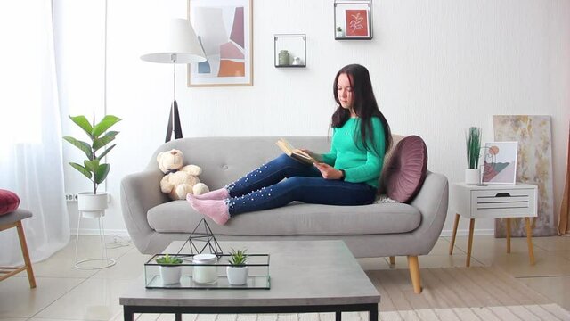 young brunette girl in jeans ang green shirt is sitting casual on the gray sofa and reading the book in light room at home in the morning. lifestyle concept, free space
