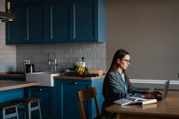 White brunette woman in eyeglasses working with laptop at home kitchen