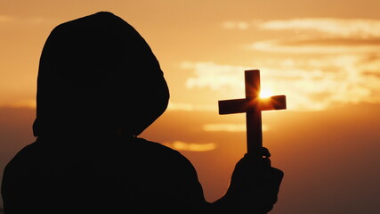 A monk in a hood with a crucifix in his hands stands against the backdrop of a dramatic sky at...