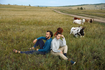Fototapeta na wymiar Hipster couple of bearded young man and girl with dreadlocks posing in the fields with cows on the background. Human and nature