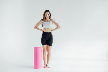 Full length portrait of a smiling sports woman with yoga mat walking over gray background. Looking away