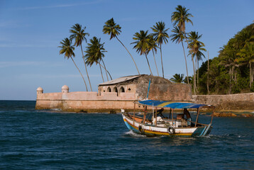 Rustic wooden fishing boat in front of the Morro de São Paulo Fortress on a sunny day. Blue sky, palm trees and forest.