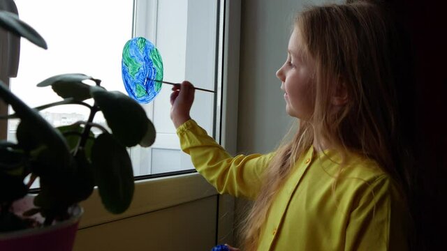 Cute little girl painting planet on window at home 4K. Happy Earth Day April 22 greeting message. Creative family leisure lockdown new reality. Ecology Saving environment conscious consumption concept