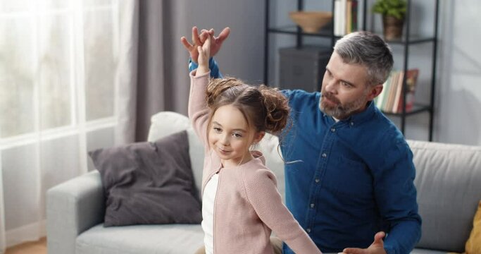 Dad and little daughter spend free time together at home on couch watching TV and dancing to melodies holding hands.