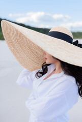 Close up of young brunette with a big hat stands on the sand. Сoncept of recreation on the beach