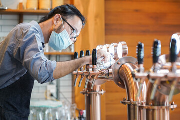 Waiter or bartender in apron and protection mask cleaning beer taps on the counter in pub and...