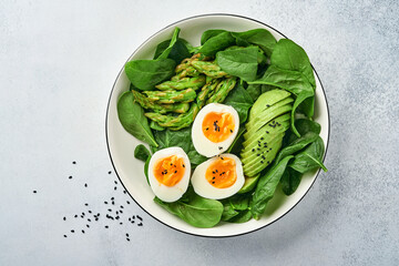 Fresh vegetable salad with avocado, asparagus, crumpled eggs with black sesame seeds and spinach on plate on light slate, stone or concrete background. Balanced lunch in bowl. Top view. Mock up.