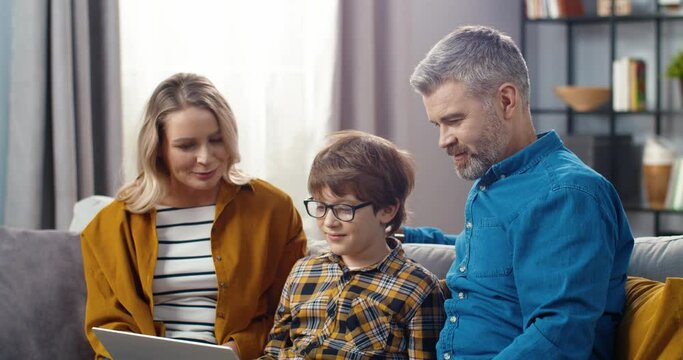 Happy mom dad and son sitting at home on couch and spending free time talking to relatives on video call using laptop.