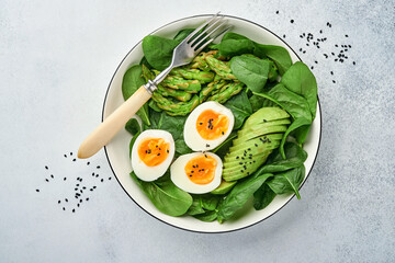 Fresh vegetable salad with avocado, asparagus, crumpled eggs with black sesame seeds and spinach on plate on light slate, stone or concrete background. Balanced lunch in bowl. Top view. Mock up.