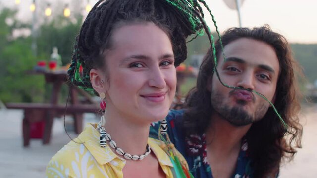 Happy Caucasian woman looking at camera and smiling while sitting on lake pier with cheerful middle eastern man holding her dreadlock as mustache
