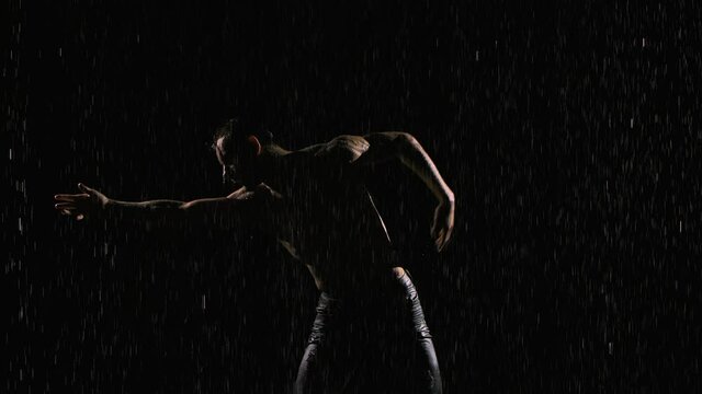 Man with naked torso moves and dances sexually. Wet athletic body is covered with shiny water droplets. Silhouette on black background under streams of rain with studio light. Close up. Slow motion.
