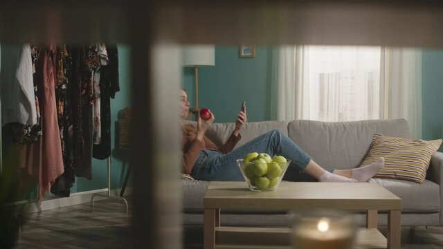 Beautiful girl in blue jeans and brown sweater lies on beige sofa on background of window, biting a red apple and and speaks via video link.