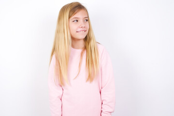 beautiful caucasian little girl wearing pink hoodie over white background with thoughtful expression, looks away keeps hands down bitting his lip thinks about something pleasant.