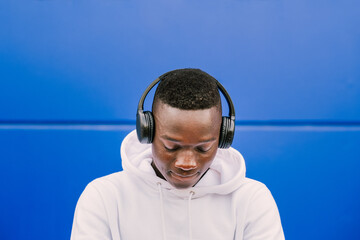 Young african black man against a blue wall wearing a white sweatshirt listening music on wireless headphones while looking down