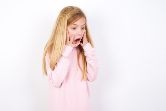 beautiful caucasian little girl wearing pink hoodie over white background with shocked facial expression, holding hands near face, screaming and looking sideways at something amazing.