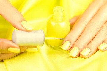 Female hands with yellow manicure on a yellow silk background. Woman painting her nails with yellow nail varnish.