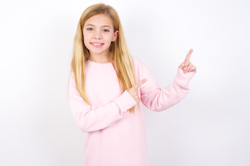 Positive beautiful caucasian little girl wearing pink hoodie over white background with beaming smile pointing with two fingers and looking on empty copy space. Advertisement concept.