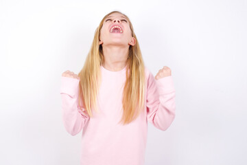 beautiful caucasian little girl wearing pink hoodie over white background looks with excitement up, keeps hands raised, notices something unexpected.